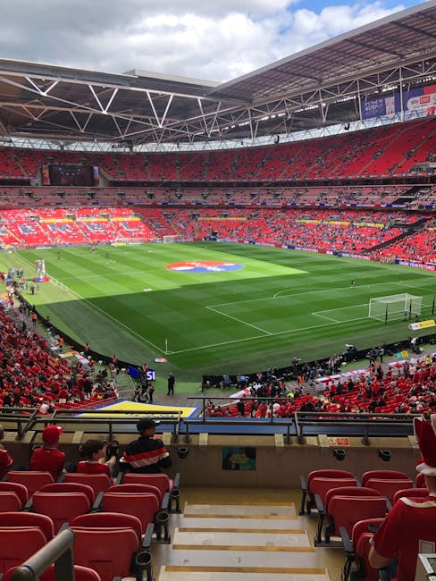 A wide view of Twickenham Stadium's indoor seating area and the central pitch during daytime. The seating sections are predominantly red with some green and white signage visible. The interior roof st